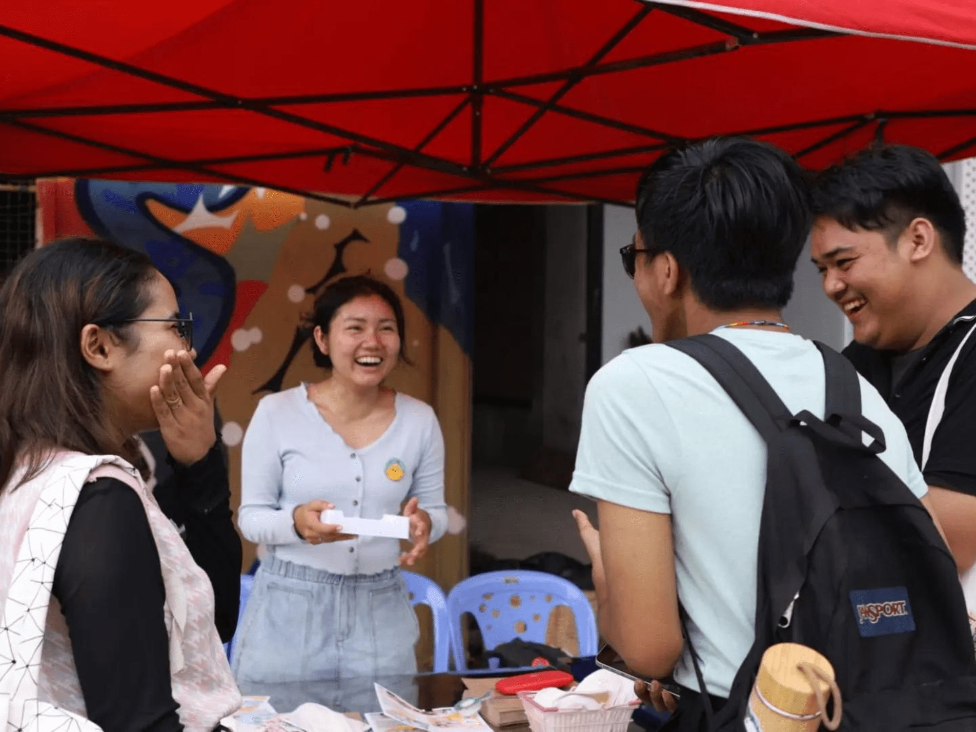 A young woman laughs at her booth at the Berk Chet Festival with three young people visiting, who are also laughing.