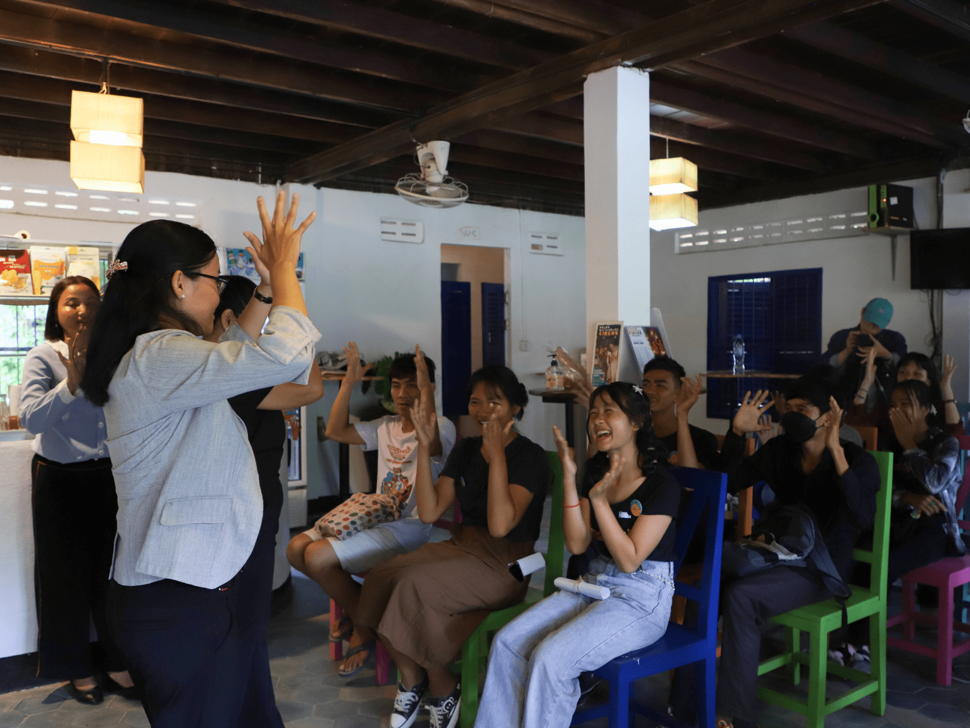 A group of people sit in rows in a room smiling and making the gesture for clapping in Cambodian Sign Language, following a leader's example at the front of the room.