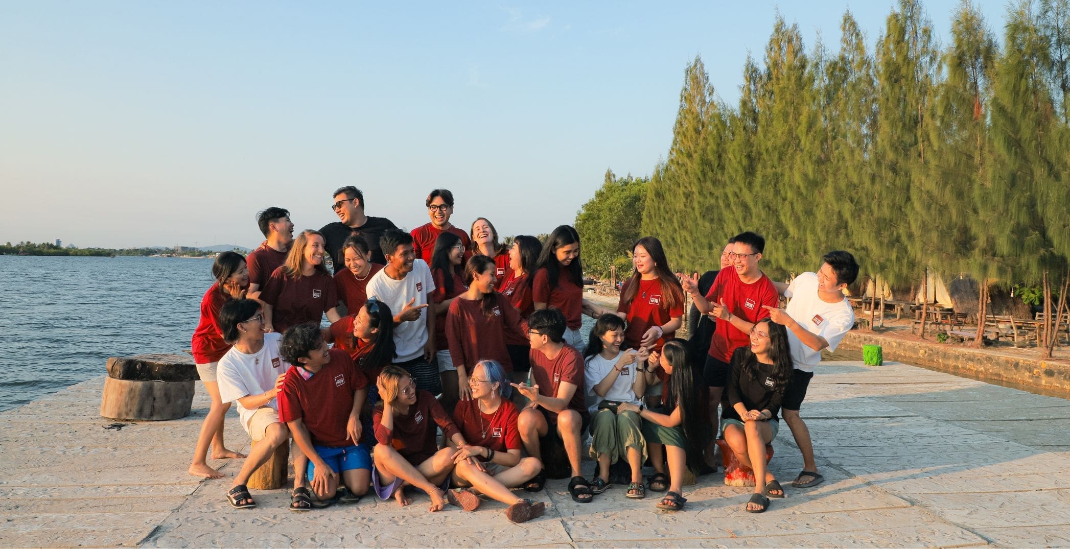 A group of around 20 Impact Hub Phnom Penh staff members make silly poses in a group photo wearing their Impact Hub t-shirts. In the background is a river in Kampot and green trees.