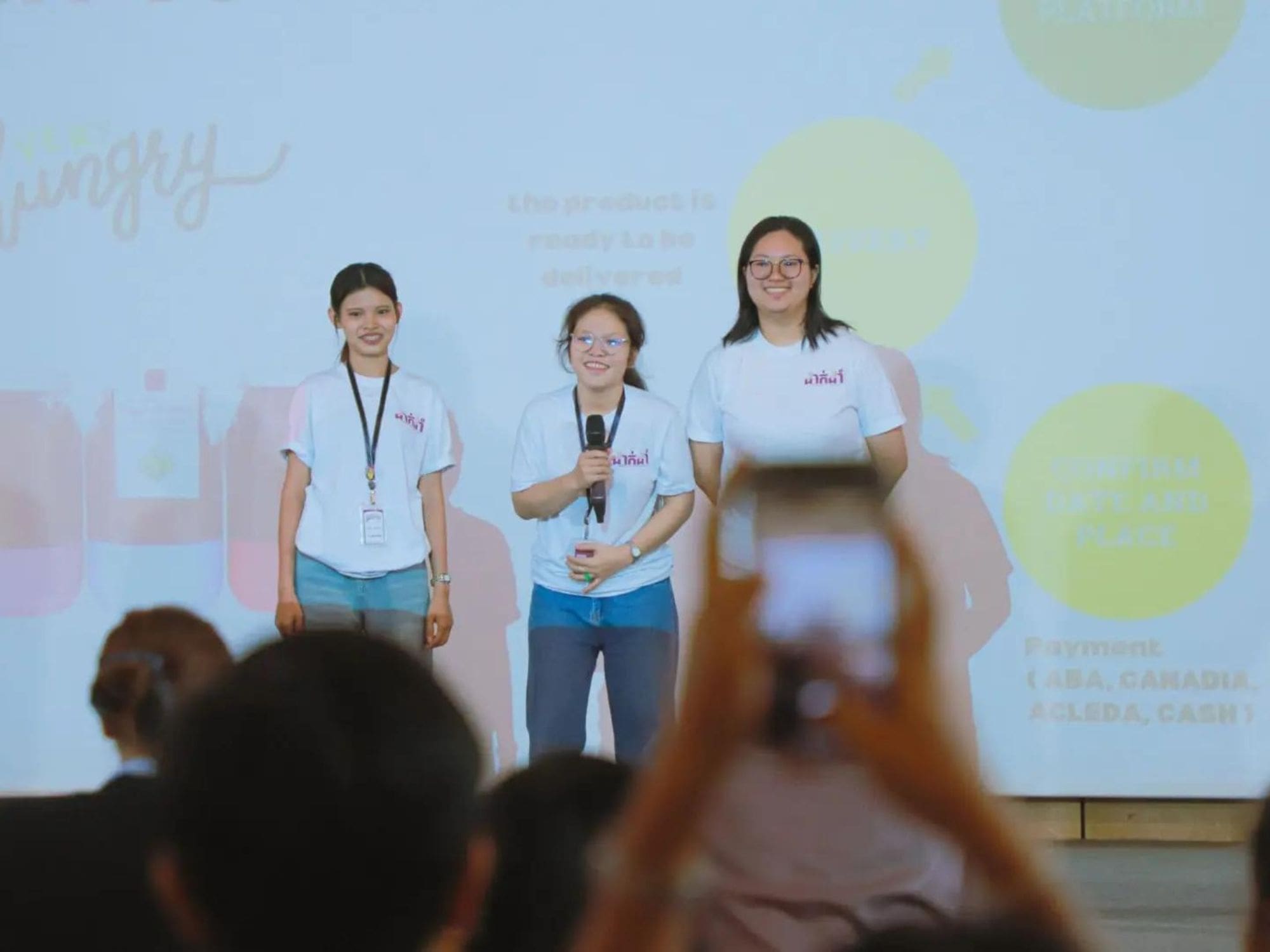 Three young women stand on a stage smiling and presenting their ideas at the DakDam hackathon.