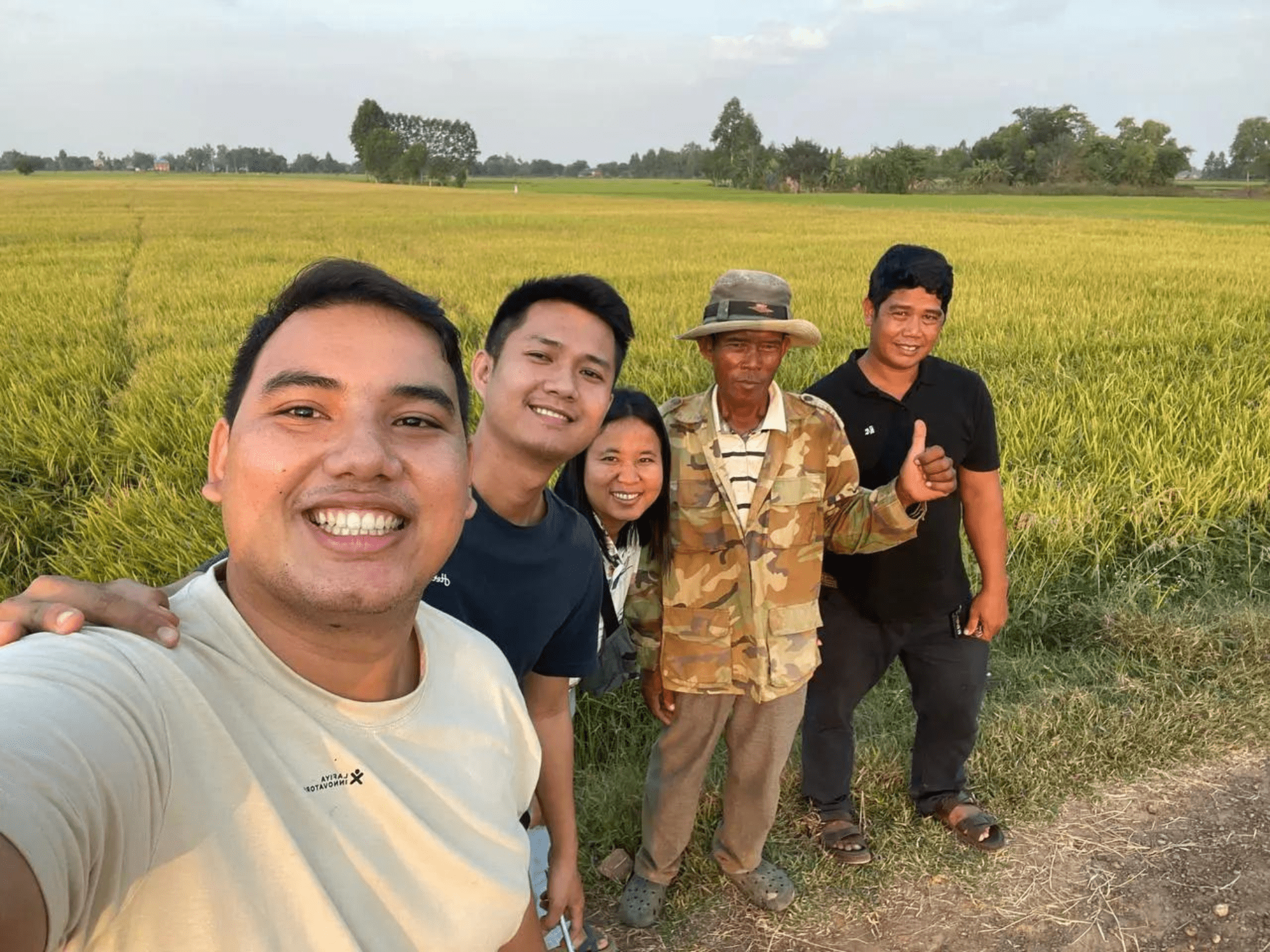 Three team members from Impact Hub Phnom Penh and two farmers stand smiling in front of a field of rice in rural Cambodia and take a selfie