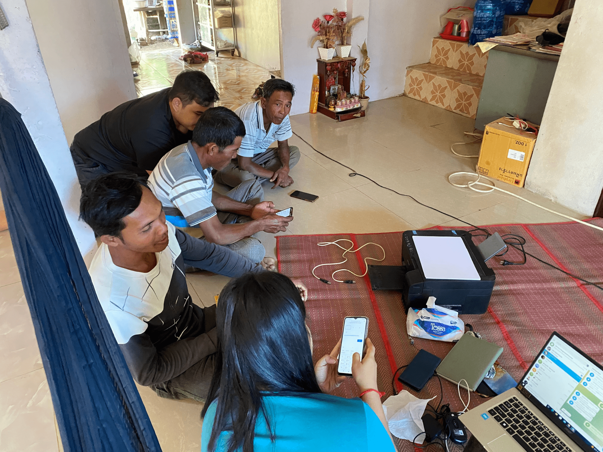 A group of people sit on a floor looking together at a few laptop and smartphone screens during an Impact Hub Phnom Penh mentoring session