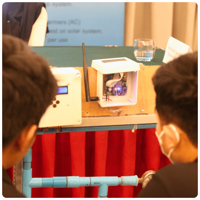 A small piece of equipment on display at a SAAMBAT Accelerator Demo Day, framed by the back of two people's heads.