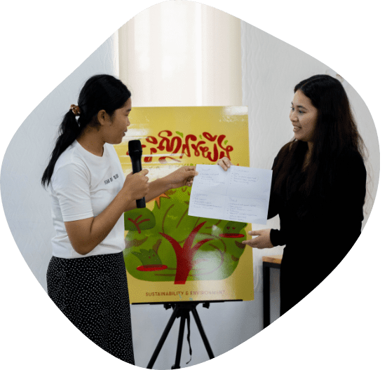 Two young Cambodian women stand in front of an illustrated poster of a tree holding up a paper and speaking into a microphone.