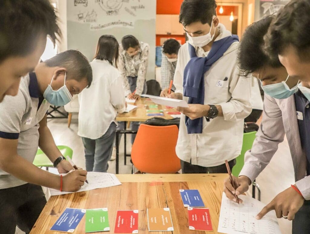 Two groups of people huddle around two tables writing on papers and re-arranging colored cards.