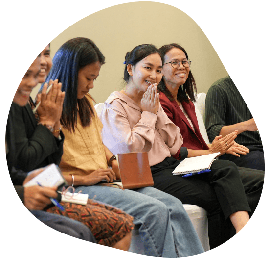 a group of women seated in a row, engaged in a discussion or event. One woman in the center, wearing a light pink blouse, is smiling and holding her hands together in a gesture of gratitude or greeting. To her right, a woman with glasses and a maroon blazer is smiling, holding a notebook, while others around them are also smiling or clapping. The atmosphere appears warm and engaging, with the participants sitting close together, suggesting an interactive or collaborative event.