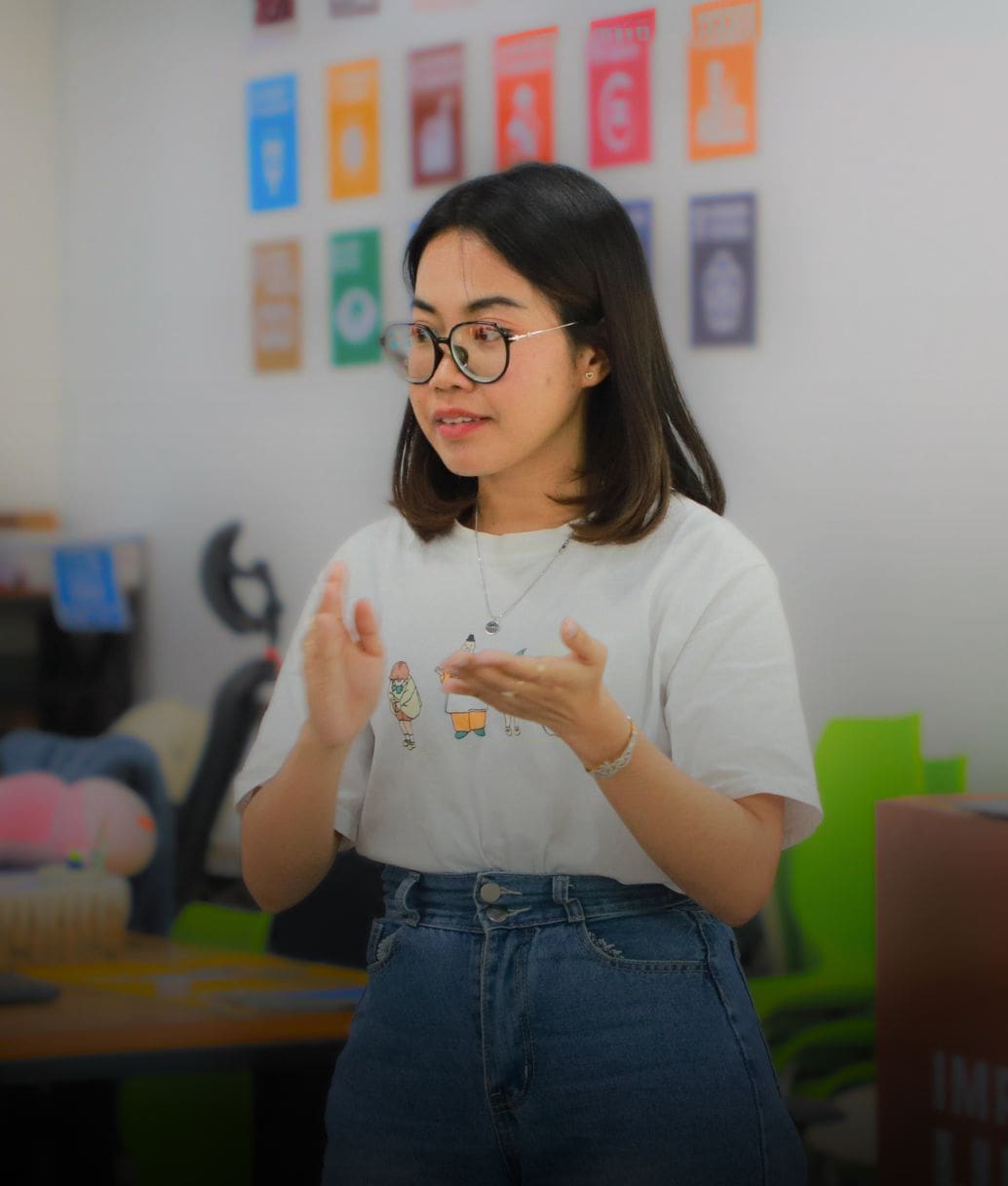 A young adult stands in front of a wall with SDG icons speaking and gesturing with her hands.