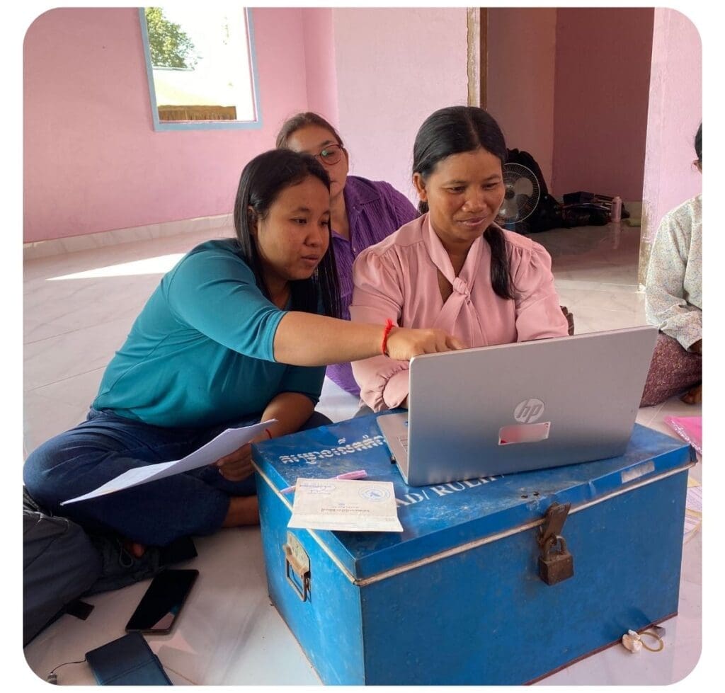 An Impact Hub Phnom Penh mentor sits on the floor with two entrepreneurs in rural Cambodia leaning over a laptop and pointing.