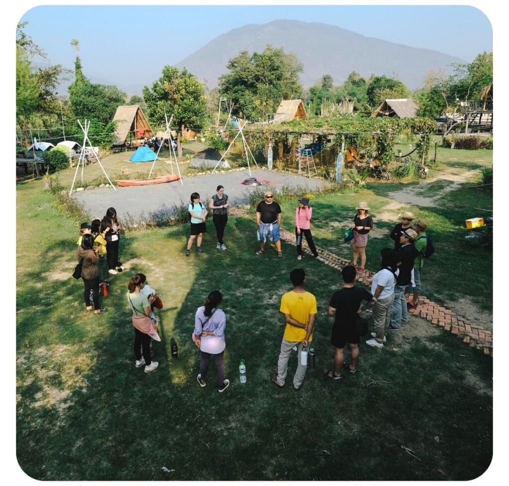 Around 15 alumni entrepreneurs of Impact Hub Phnom Penh stand in a large circle outside. It's a zoomed out photo with a campsite and mountains seen in the background.