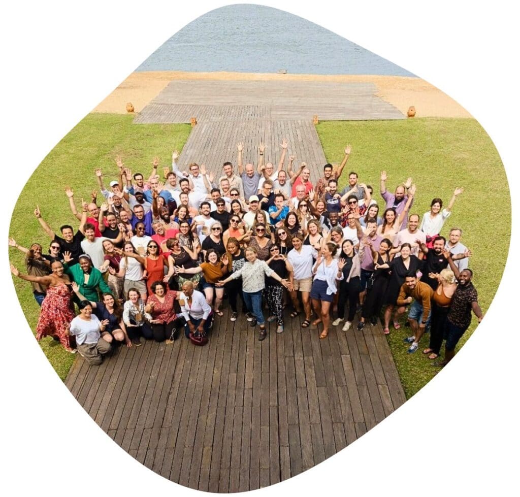 Over fifty people from Impact Hubs all over the world pose for a group photo outside, smiling and waving their arms in the air.