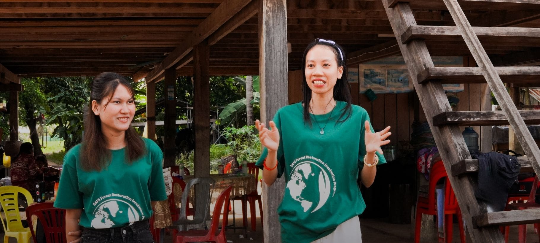 Two young Cambodian women stand in front of a traditional Khmer house. One is speaking and gesturing with her hands while the other looks at her.