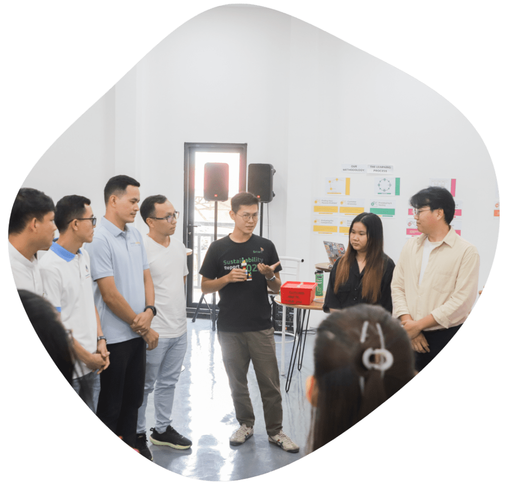 A group of around 7 people stand in a semi circle in a bright room as a man in the middle speaks.