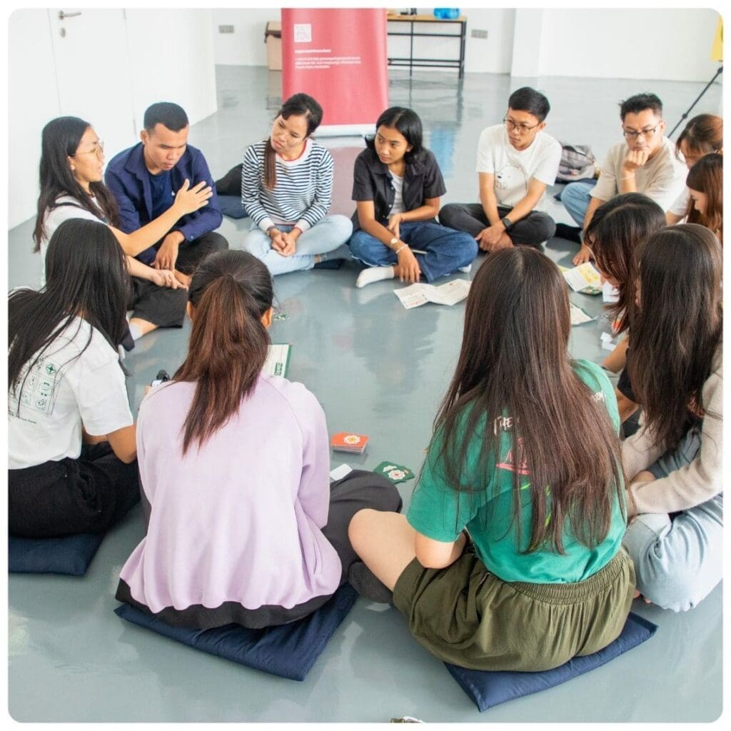 A group of around 10 young people sit on cushions on the floor in a circle listening to a woman speak at Impact Hub Phnom Penh's event space.