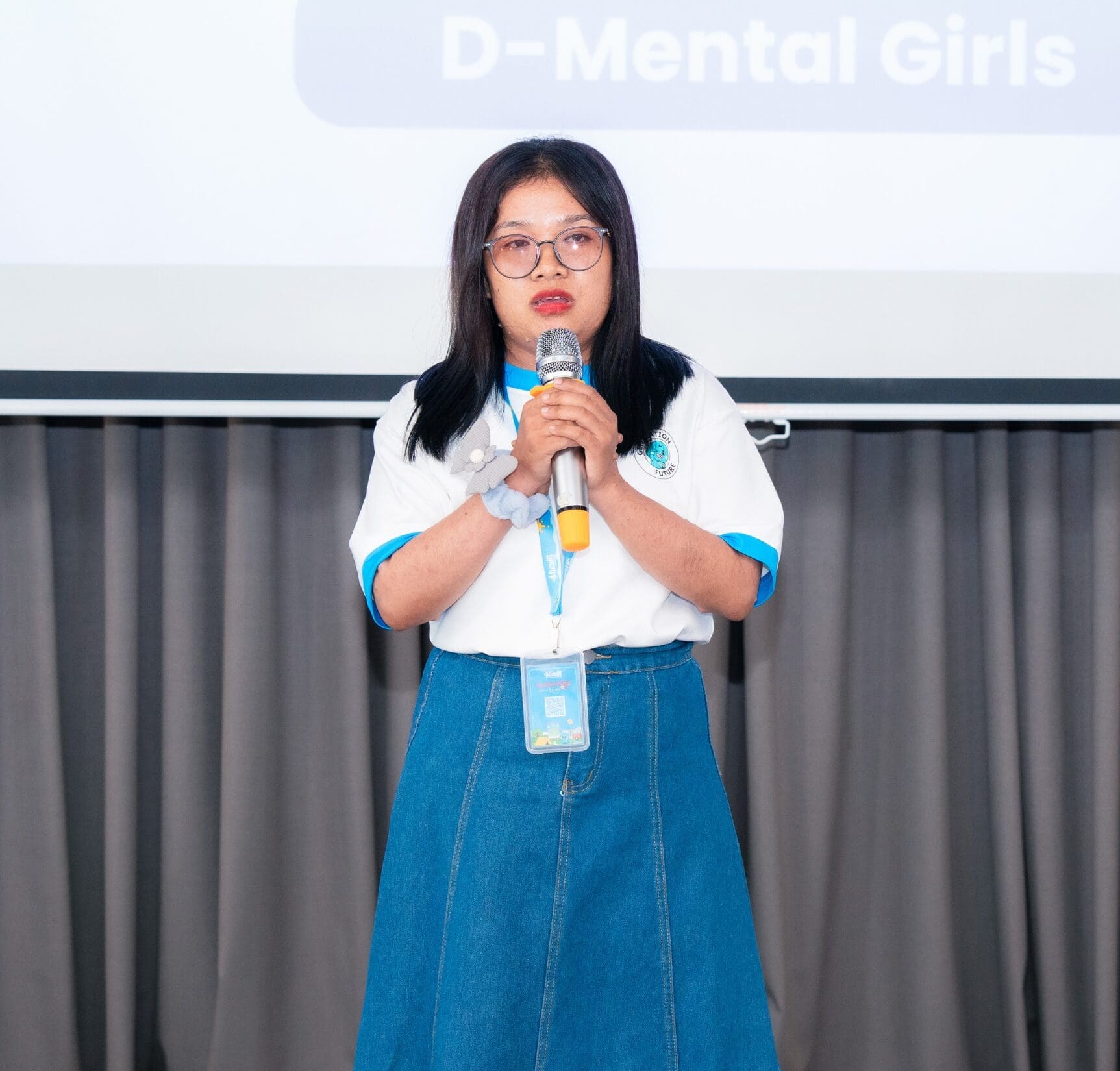 A person stands holding a microphone, wearing glasses, a white and blue shirt, and a blue skirt. A screen behind them displays the text "D-Mental Girls."