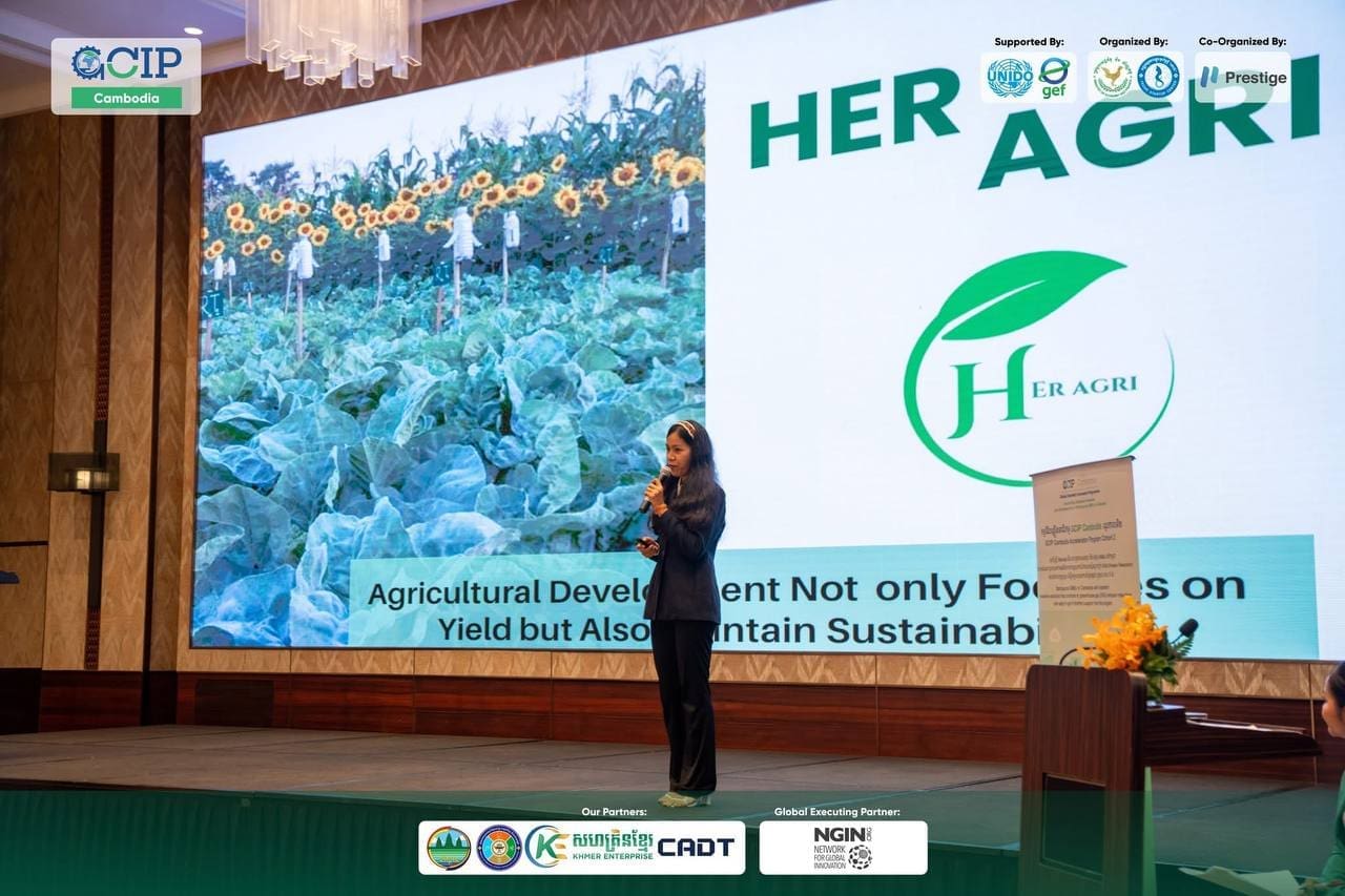 A woman speaks onstage at a conference. Behind her, a large screen displays an image of crops and the text "HER AGRI," emphasizing sustainable agriculture.