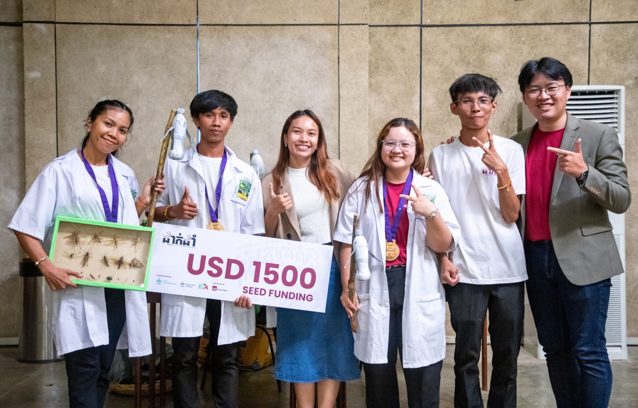 A group of six smiling people pose with a USD 1500 seed funding sign. Two in lab coats hold an insect display. The mood is celebratory and proud.