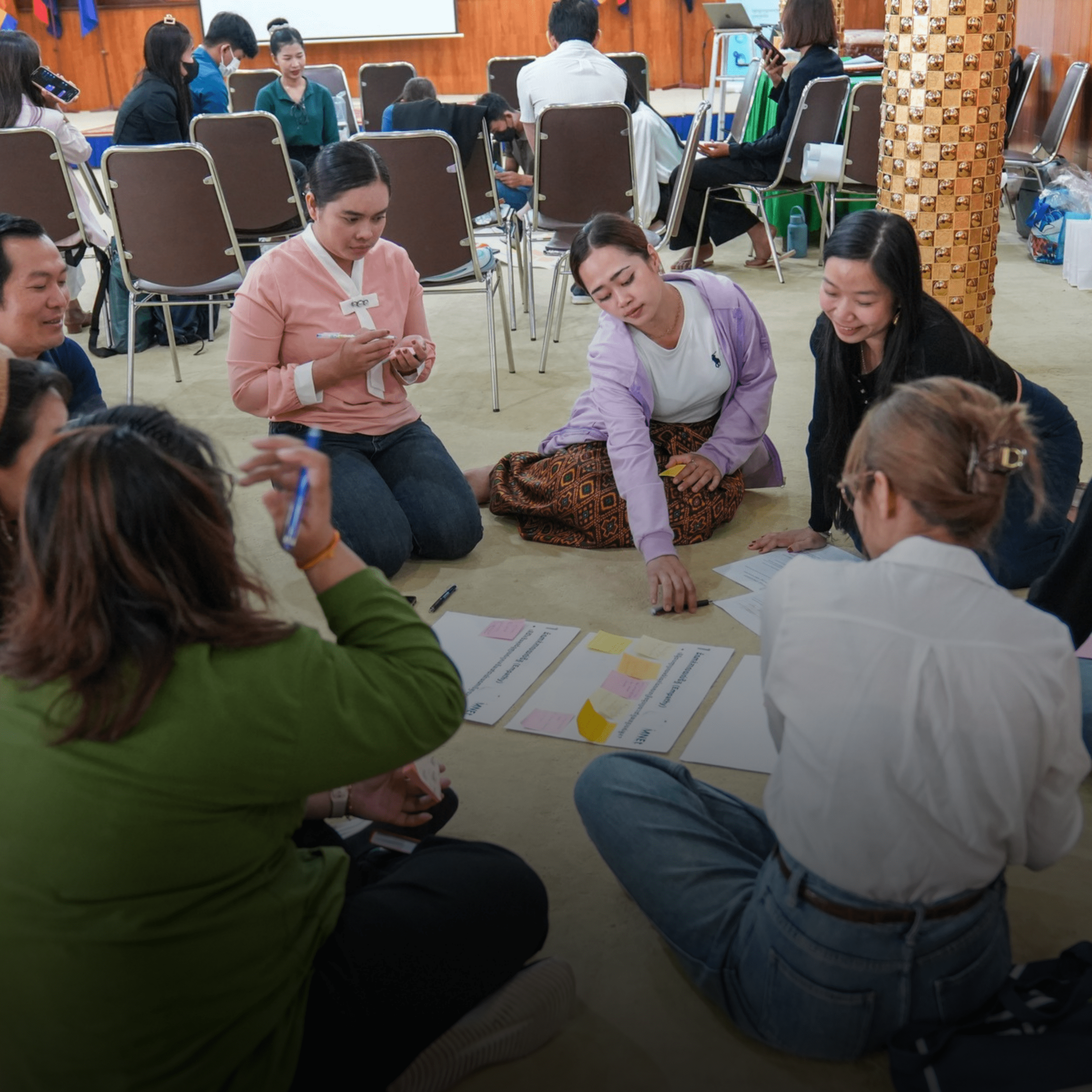 A group of people sitting on the floor in a circle, engaged in a collaborative activity with papers and sticky notes in a relaxed, focused setting.