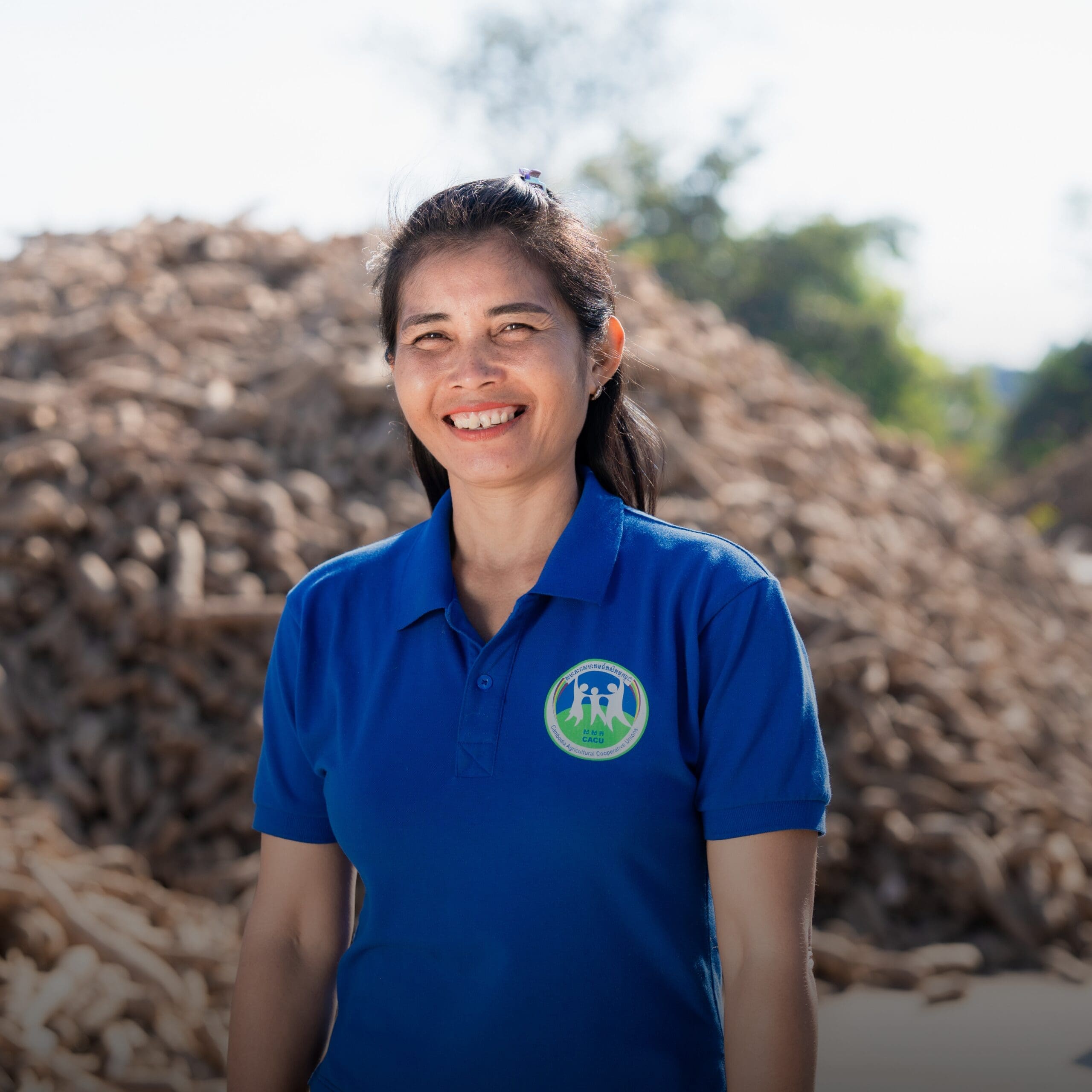 Woman in a blue shirt, smiling warmly, stands in front of a large pile of tubers outdoors, with trees and a bright sky in the background.
