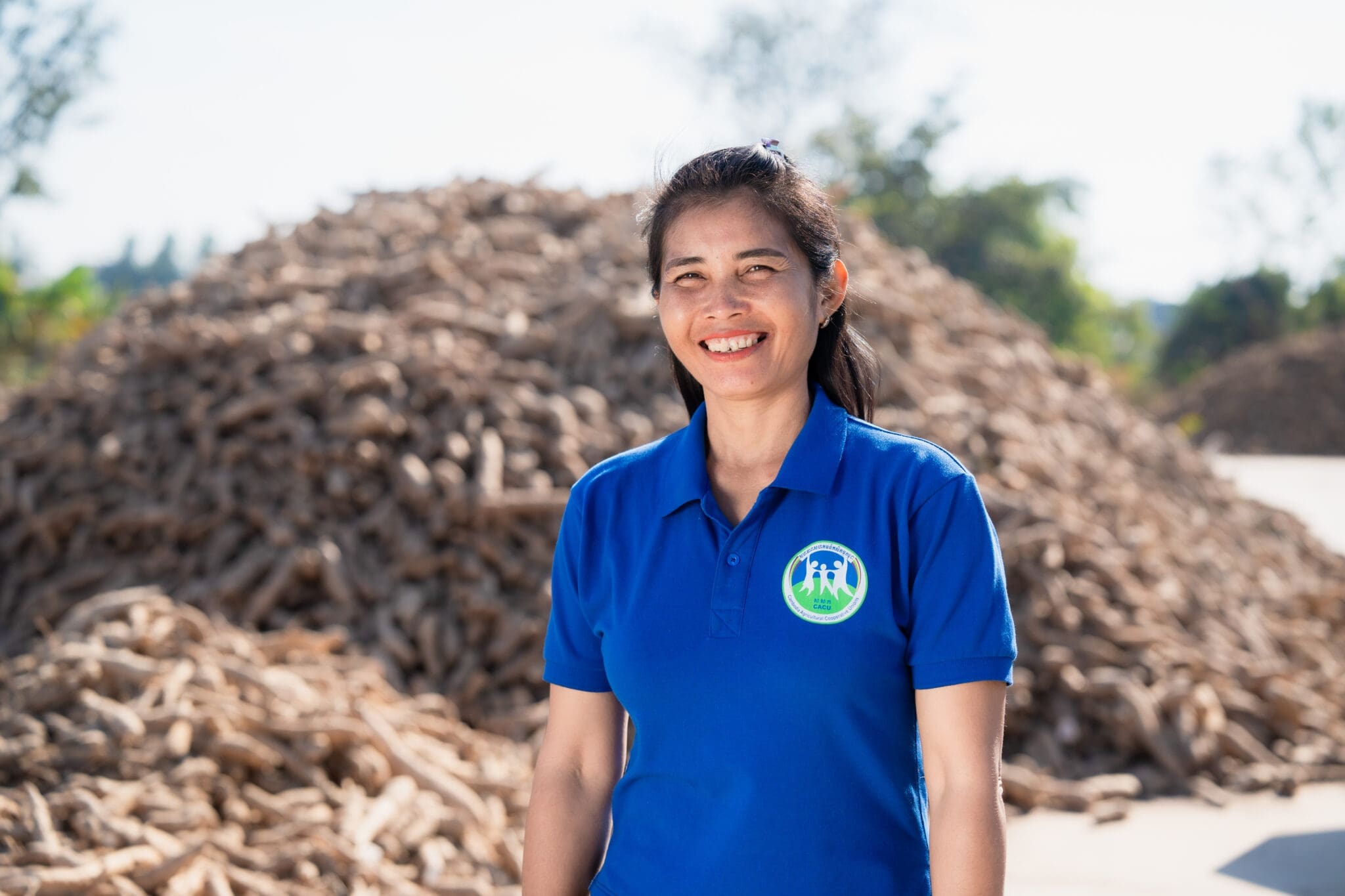 A woman in a blue polo shirt with a logo stands smiling in front of a large pile of dried roots outdoors, under a clear sky. The scene conveys positivity.