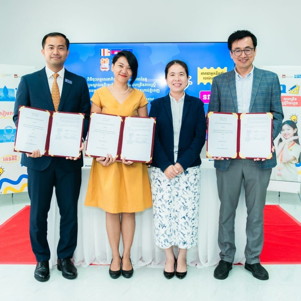 Four people stand in a bright room holding framed certificates, smiling. A digital display and banners in the background suggest a formal event.
