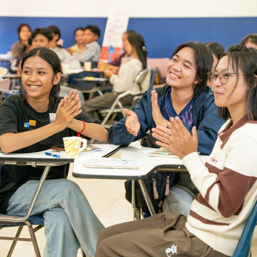 A group of young students sits around tables in a classroom setting, smiling and clapping. The atmosphere is lively and positive, suggesting engagement and teamwork.