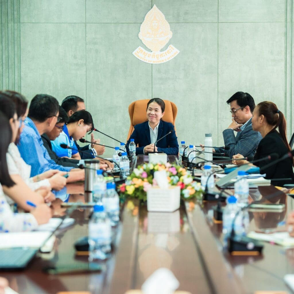 Meeting room with a central person smiling, surrounded by diverse individuals discussing. A flower arrangement and water bottles on the table add color.