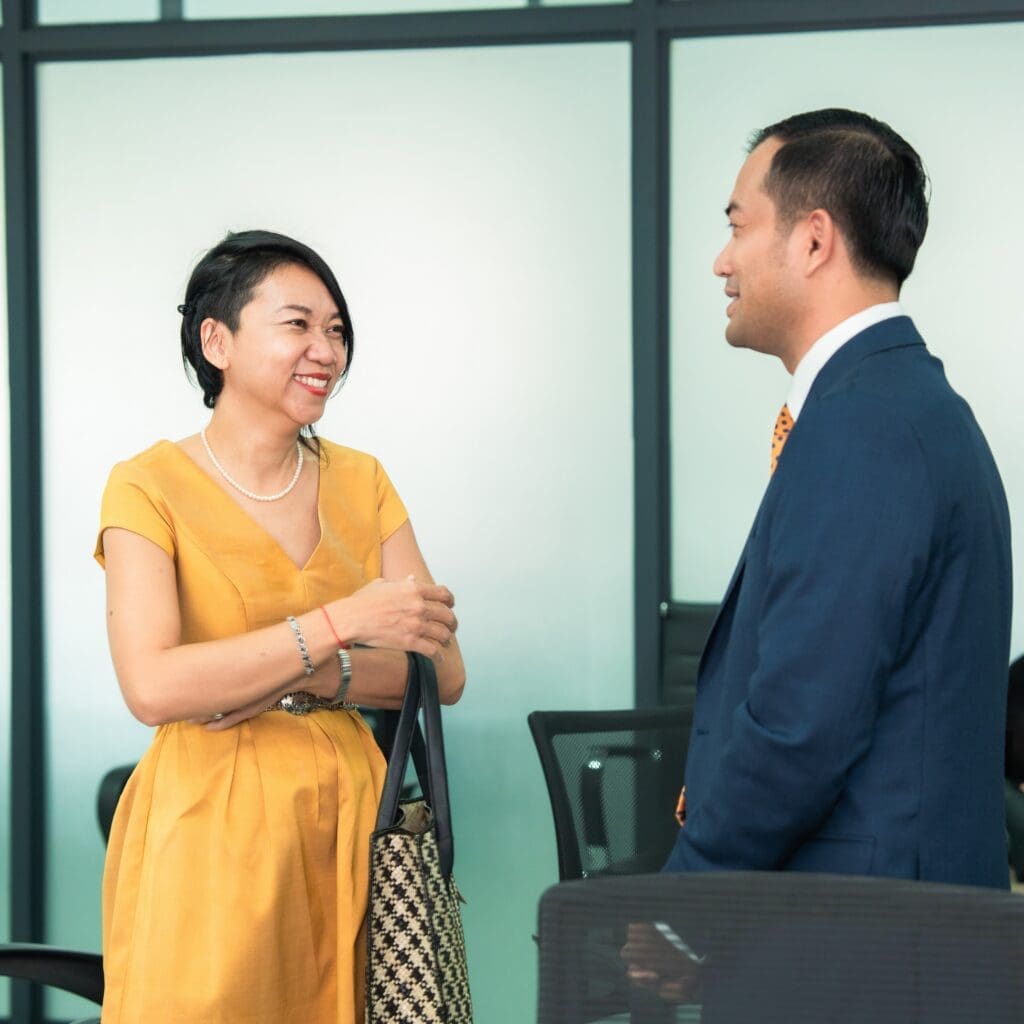 A woman in a yellow dress and a man in a blue suit stand in an office, smiling and conversing, creating a warm and professional atmosphere.