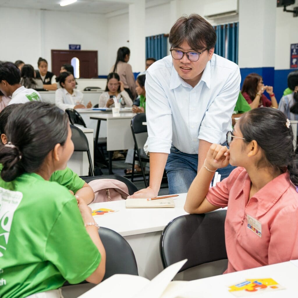 A teacher in a blue shirt engages with students in a classroom, leaning over a desk. The atmosphere is lively, with students listening attentively.