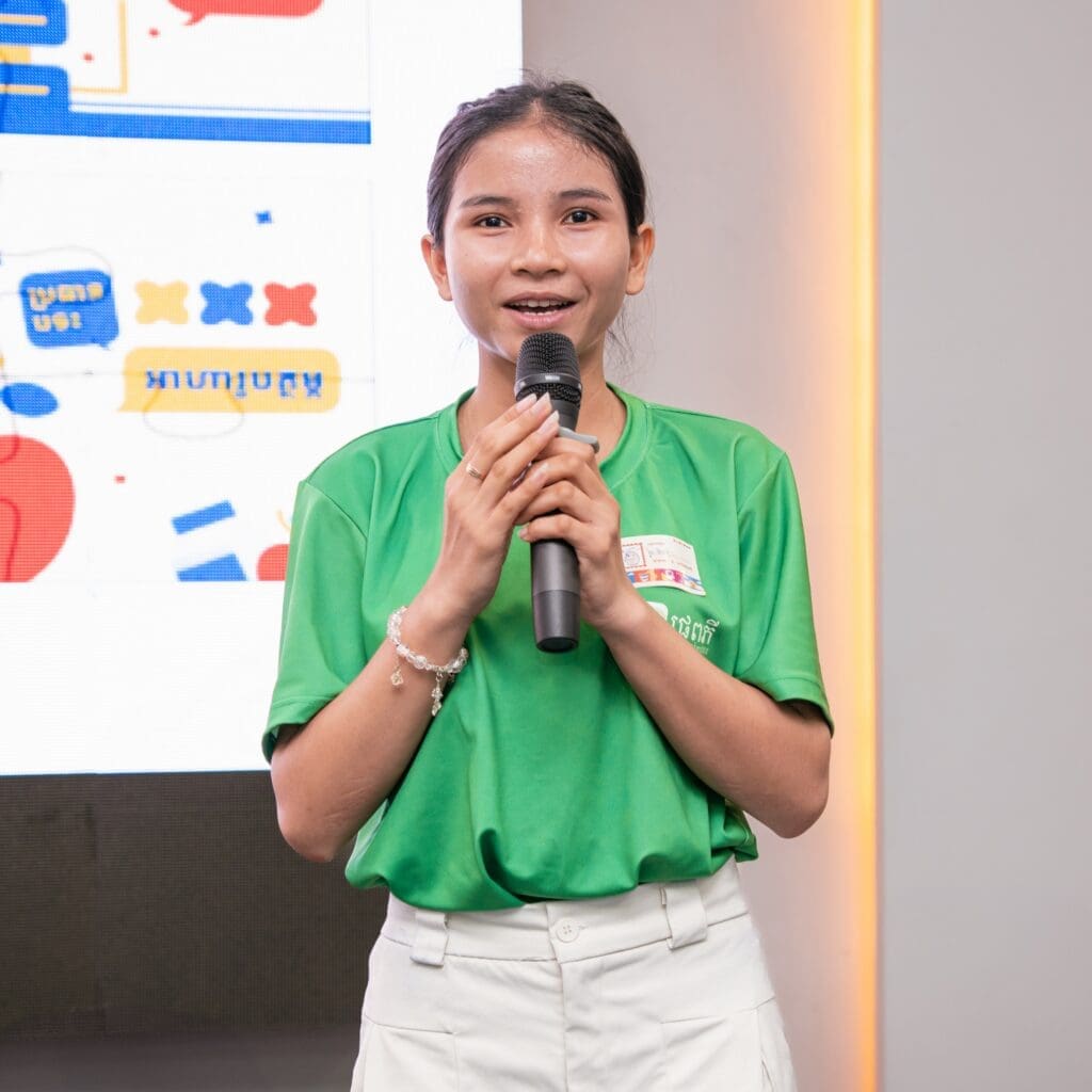 A young woman in a green shirt holds a microphone, speaking confidently. Behind her is a bright digital screen with colorful graphics and text.