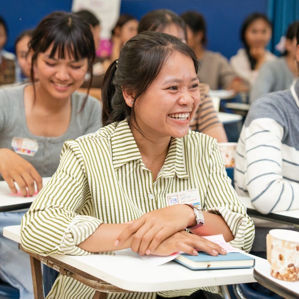 A woman in a striped shirt sits at a desk, smiling and looking engaged in a classroom setting. Other students are seen in the background, also smiling.