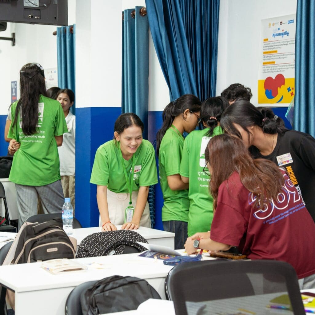A group of young people in bright green and red shirts collaborate in a classroom. They appear engaged and cheerful, interacting around desks with books and backpacks.