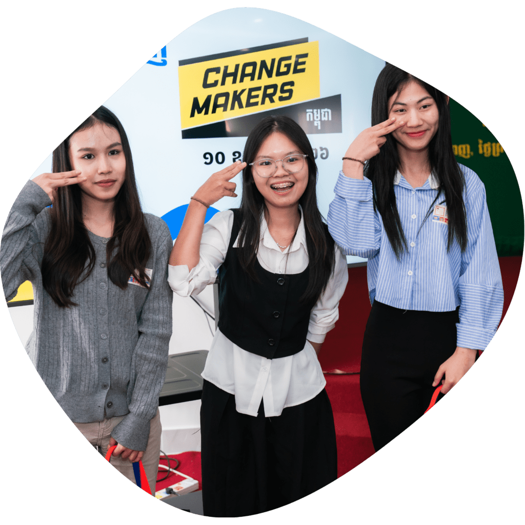 Three women stand smiling, making a peace sign near their faces. Behind them, a sign reads "Change Makers." The mood is cheerful and empowering.